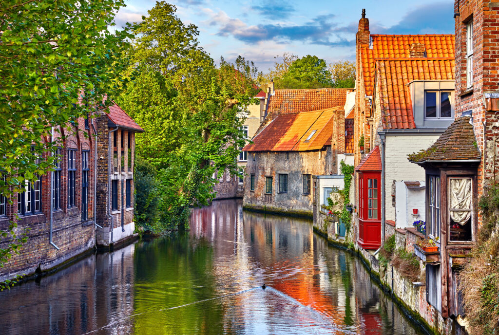 Bruges, Belgium. Medieval ancient houses made of old bricks at water channel with boats in old town. Summer sunset with sunshine and green trees. Picturesque landscape.